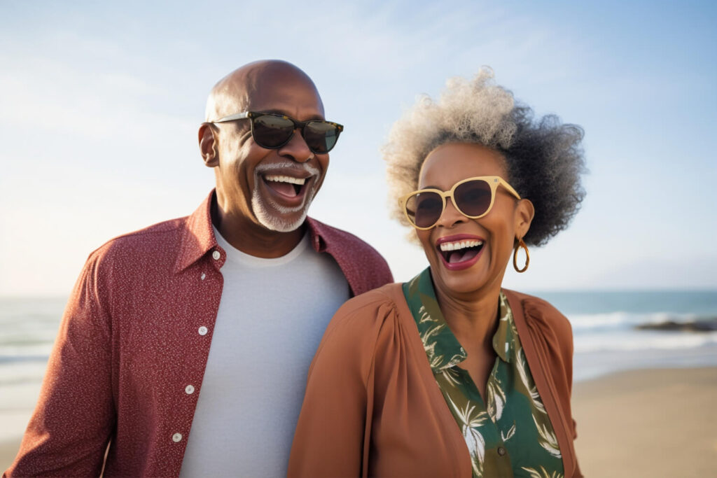 Casal sênior negro sorrindo e rindo muito na praia, usando óculos de sol, ilustrando a alegria de uma reabilitação oral completa em Sergipe.